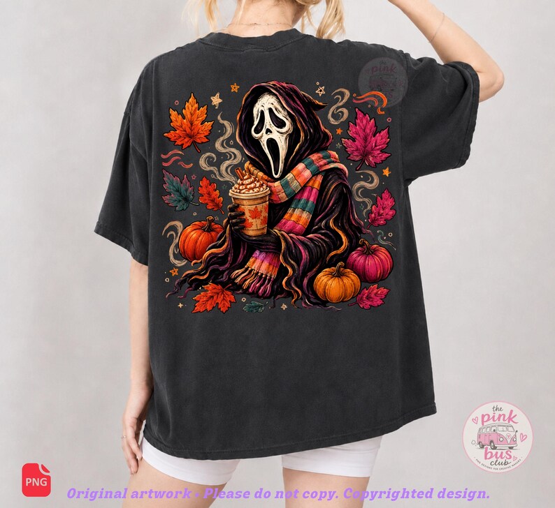 May include: A charcoal black t-shirt featuring a graphic print of a hooded figure with a white mask holding a coffee cup, surrounded by orange and pink autumn leaves, pumpkins, and stars. The design includes a colorful striped scarf and swirls.