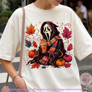 May include: An off-white t-shirt featuring a graphic of a hooded figure with a white mask holding a coffee cup, surrounded by orange and pink pumpkins and autumn leaves. The figure wears a colorful striped scarf, and steam rises from the cup.