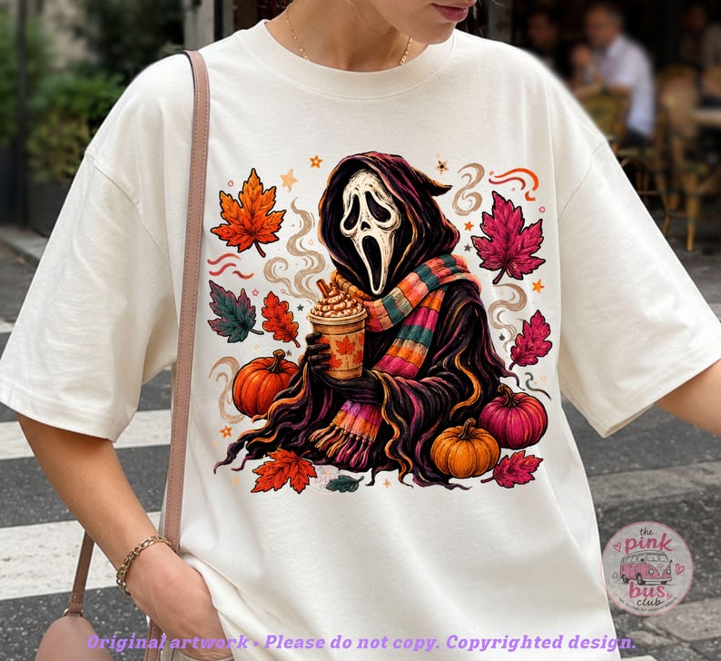 May include: An off-white t-shirt featuring a graphic of a hooded figure with a white mask holding a coffee cup, surrounded by orange and pink pumpkins and autumn leaves. The figure wears a colorful striped scarf, and steam rises from the cup.