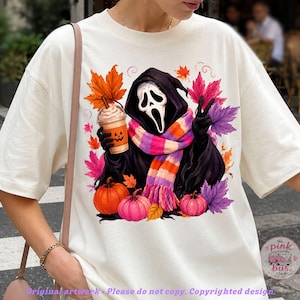 May include: An off-white t-shirt featuring a graphic of a hooded figure with a white mask, wearing a pink, orange, and purple striped scarf. The figure holds a pumpkin-faced coffee cup and makes a peace sign, surrounded by colorful autumn leaves.