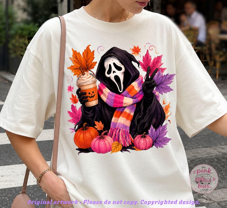 May include: An off-white t-shirt featuring a graphic of a hooded figure with a white mask, wearing a pink, orange, and purple striped scarf. The figure holds a pumpkin-faced coffee cup and makes a peace sign, surrounded by colorful autumn leaves.