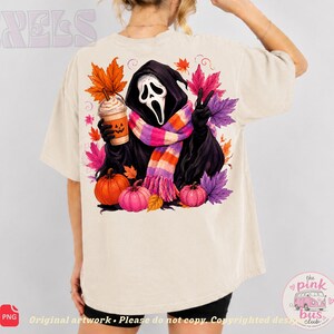 May include: A beige t-shirt featuring a graphic of a hooded figure with a white mask, wearing a pink, orange, and purple striped scarf. The figure holds a pumpkin-faced coffee cup, surrounded by colorful autumn leaves and two pumpkins.