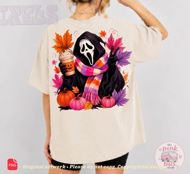 May include: A beige t-shirt featuring a graphic of a hooded figure with a white mask, wearing a pink, orange, and purple striped scarf. The figure holds a pumpkin-faced coffee cup, surrounded by colorful autumn leaves and two pumpkins.