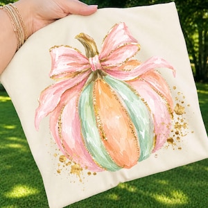 May include: A cream-colored fabric item featuring a watercolor-style illustration of a pumpkin. The pumpkin is painted in shades of pink, mint green, and orange, accented with gold glitter outlines and a large pink bow tied around the stem.