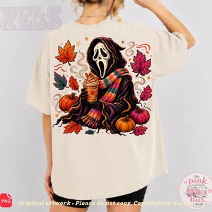 May include: A beige t-shirt featuring a back graphic of a hooded figure with a white mask, wearing a colorful striped scarf and holding a coffee cup. The design includes orange and pink autumn leaves, two pumpkins, and swirling smoke elements.