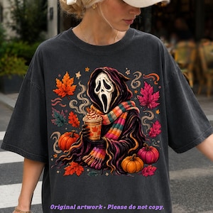 May include: A charcoal gray t-shirt featuring a graphic of a hooded figure with a white mask holding a coffee cup. The design includes a colorful striped scarf, orange and pink pumpkins, and scattered autumn leaves in shades of red and orange.