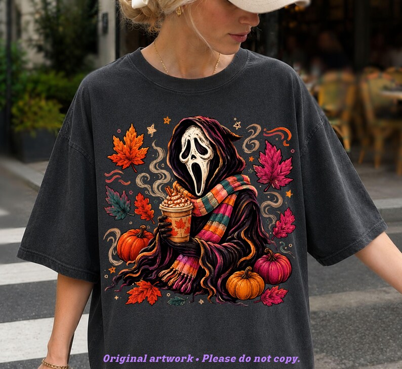 May include: A charcoal gray t-shirt featuring a graphic of a hooded figure with a white mask holding a coffee cup. The design includes a colorful striped scarf, orange and pink pumpkins, and scattered autumn leaves in shades of red and orange.