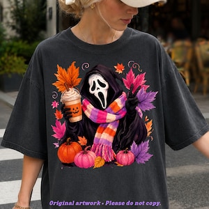 May include: A charcoal gray t-shirt featuring a graphic of a hooded figure with a white mask, wearing a striped scarf, holding a pumpkin-faced coffee cup, and making a peace sign. The design includes orange, pink, and purple autumn leaves and pumpkins.