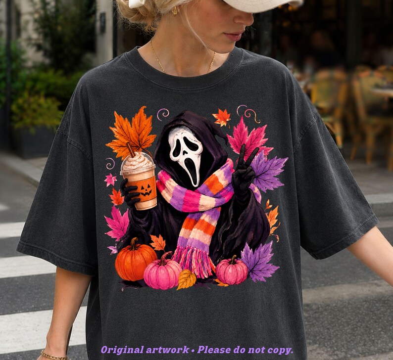May include: A charcoal gray t-shirt featuring a graphic of a hooded figure with a white mask, wearing a striped scarf, holding a pumpkin-faced coffee cup, and making a peace sign. The design includes orange, pink, and purple autumn leaves and pumpkins.