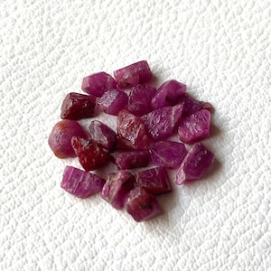 Puede incluir: Un conjunto de piedras preciosas de rubí en bruto, en tonos rojos y rosas. Las piedras sin cortar se muestran sobre una superficie blanca, resaltando sus formas y texturas naturales e irregulares. Ideal para la fabricación de joyas.