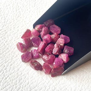 Puede incluir: Una colección de gemas de rubí en bruto sin cortar. Los rubíes en bruto varían en tamaño y forma, mostrando un tono rojo-rosa intenso. Se encuentran sobre una superficie blanca, algunos en un recogedor negro.