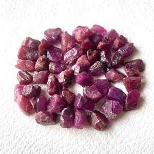 Puede incluir: Una colección de pequeñas piedras preciosas de rubí en bruto y sin pulir en varios tonos de rosa intenso y rojo. Cada piedra presenta una forma irregular y dentada con una superficie cristalina texturizada y variaciones naturales.