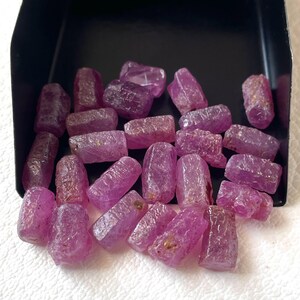 Puede incluir: Una colección de pequeñas piedras preciosas de rubí en bruto y sin pulir con un tono rosa rojizo vibrante y superficies irregulares y texturizadas. Estos cristales opacos sueltos están contenidos dentro de una pala de metal negro.
