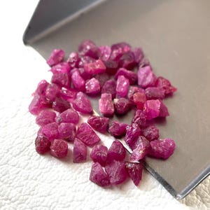 Könnte beinhalten: Eine Ansammlung kleiner, roher, dunkelrosa bis magentafarbener Rubin-Edelsteine, die auf einer silbernen Metallschaufel verstreut sind. Die Steine haben unregelmäßige, facettierte und raue Oberflächen und zeigen verschiedene Rot- und Rosatöne.