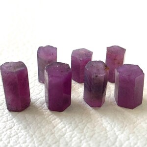 Puede incluir: Un conjunto de siete pequeños cristales de rubí en bruto de forma hexagonal. Cada piedra presenta un tono magenta rosado intenso con variaciones naturales en la intensidad del color y la textura de la superficie.