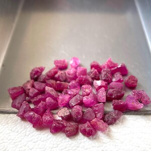 Könnte beinhalten: Eine Ansammlung kleiner, roher, tiefrosa bis magentafarbener Edelsteinsplitter, die in einer metallisch silberfarbenen Schaufel liegen. Die Steine haben unregelmäßige, gezackte Formen und facettierte Oberflächen, die das Licht reflektieren und unterschiedliche Grade an Transparenz und Textur aufweisen.