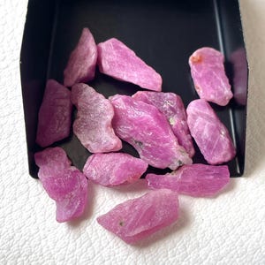Puede incluir: Una colección de piedras preciosas de rubí en bruto, sin pulir, esparcidas dentro de una pala de metal negro. Las piedras presentan formas irregulares y dentadas con texturas cristalinas naturales y tonos variados de magenta vibrante y rosa.
