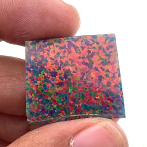 Puede incluir: Una piedra preciosa de ópalo de forma cuadrada que presenta un vibrante juego de colores con motas dispersas de rojo brillante, naranja, amarillo, verde y azul sobre una base translúcida. La superficie plana y pulida muestra un patrón denso.