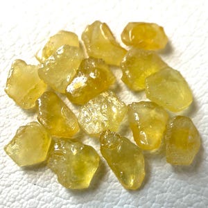 Puede incluir: Una colección de pequeñas piedras preciosas amarillas en bruto con formas irregulares y angulares y superficies translúcidas. Estos fragmentos minerales naturales exhiben varios tonos de amarillo brillante y oro, con bordes facetados.