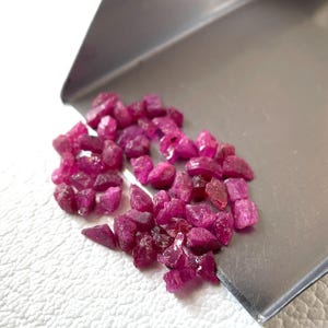Könnte beinhalten: Eine Ansammlung kleiner, roher, tiefrosa Rubinedelsteine, die auf einer silbernen Metallschaufel verstreut sind. Die Steine haben unregelmäßige, gezackte Formen und eine durchscheinende, kristalline Textur, die mit der glatten Oberfläche kontrastiert.