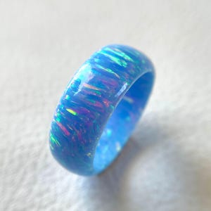 Puede incluir: Un anillo de ópalo azul con una banda ancha. El anillo tiene un acabado brillante y muestra colores iridiscentes de rosa, verde y morado. El anillo es talla 14.