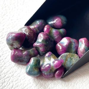 Puede incluir: Una colección de piedras pulidas que presentan una mezcla de tonos magenta vibrante, morado intenso y verde azulado. Estos especímenes minerales pulidos de formas irregulares se encuentran reunidos dentro de una pala de metal negro mate.