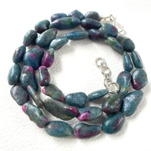 May include: A necklace composed of polished, irregular, tumbled gemstone beads in shades of deep teal, blue, and magenta. The strand features a silver-toned metal clasp and an adjustable chain link extension, creating a continuous circular arrangement.