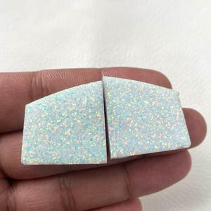 Puede incluir: Dos cabujones de ópalo blanco en forma trapezoidal que presentan un vibrante juego de colores iridiscentes con destellos azules, verdes y naranjas en toda la superficie. Estas piedras preciosas pulidas tienen superficies planas y bordes rectos.