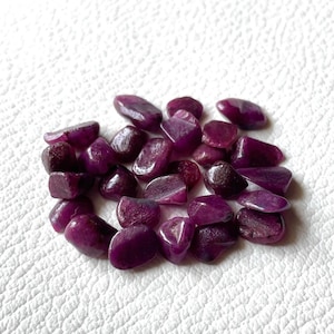 Puede incluir: Un conjunto de piedras preciosas pulidas de color púrpura oscuro. Las piedras varían en tamaño y forma, con una superficie lisa y reflectante. Las gemas están dispersas sobre un fondo blanco texturizado, creando un contraste de color y textura.