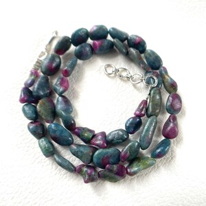 May include: A necklace composed of tumbled, irregular-shaped beads in shades of deep teal, blue-green, and magenta-pink. The strand features a silver-toned metal clasp with an adjustable chain link extension, securing the polished gemstone beads.