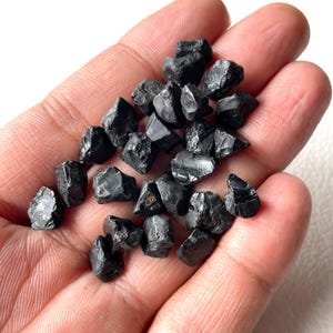 Puede incluir: Una colección de pequeñas piedras de turmalina negra en bruto con formas irregulares y angulares, que presentan una textura superficial brillante y reflectante. Estos fragmentos minerales oscuros y opacos tienen bordes afilados.