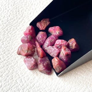 Puede incluir: Un conjunto de piedras preciosas de rubí en bruto, sin cortar. Las piedras, de diferentes tamaños y formas, muestran tonos rojos y rosados. Se presentan en una bandeja negra sobre un fondo blanco texturizado, resaltando su color natural.