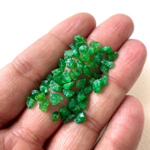 Puede incluir: Una colección de pequeños fragmentos de piedras preciosas en bruto de color verde vibrante, con bordes irregulares y dentados, y una textura cristalina translúcida. Estas piezas minerales sueltas y sin pulir varían en tamaño y forma.