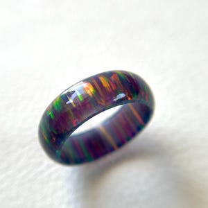 Può includere: Un anello di opale scuro e iridescente. L'anello mostra uno spettro di colori, tra cui verde, arancione e viola, nella sua superficie lucida. L'anello è posto su uno sfondo bianco.
