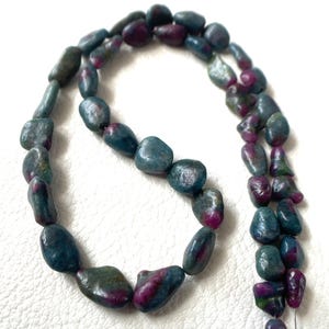 Puede incluir: Un collar de cuentas de piedras preciosas pulidas, irregulares y redondeadas, que presenta una mezcla de tonos azul verdoso profundo, turquesa y rosa magenta vibrante. Las piedras son lisas, redondeadas y varían en tamaño y forma.