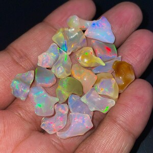 Puede incluir: Una colección de ópalos pequeños, irregulares y translúcidos que muestran un juego de colores con destellos de verde neón, rojo brillante, azul eléctrico y amarillo. Estas piedras preciosas pulidas tienen bordes suaves y redondeados, con una base blanca lechosa y vibrantes reflejos de luz internos visibles en todas las piedras.