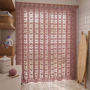 Puede incluir: Una cortina de ganchillo color rosa empolvado con paneles verticales de intrincados patrones calados y un borde inferior con flecos. La cortina cuelga de una barra de madera mediante bucles de ganchillo integrados, creando una cubierta texturizada.