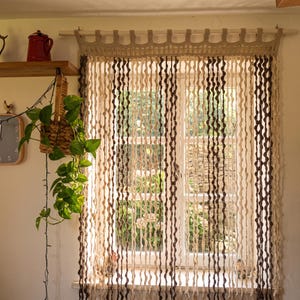 Puede incluir: Una cortina de ventana de ganchillo con hilos verticales alternos de color crema y marrón oscuro. La cortina cuelga de una barra de madera con un encabezado decorativo de ganchillo, creando una apariencia ondulada y texturizada.