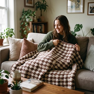 Può includere: Una coperta all'uncinetto caratterizzata da un motivo pied-de-poule ripetuto nei colori marrone scuro e crema. Il tessuto presenta un aspetto intrecciato e testurizzato con un design a griglia coerente su tutta la superficie.