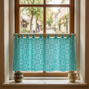 Op de afbeelding: Een turquoise gehaakt café-gordijn met een herhalend vierkant rasterpatroon. Het gordijn hangt aan een dunne koperen roede door middel van geïntegreerde lussen langs de bovenrand en vertoont een delicate, opengewerkte textuur in de stof.