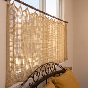 May include: A beige crochet curtain with a square grid pattern hanging from a dark brown wooden rod. The curtain features a series of loops at the top for attachment to the rod, creating vertical folds across the fabric that cover the window.