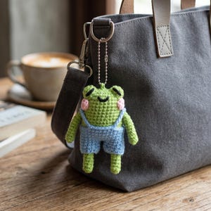 May include: A green crochet frog keyring with pink cheeks and blue dungarees hangs from a grey tote bag. The frog has a smiling face and is attached to a silver chain. A cup of coffee is in the background.
