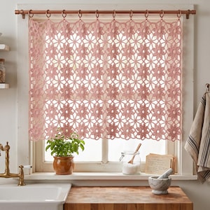 Può includere: Una tenda all'uncinetto color rosa polvere con un motivo floreale ripetuto. La tenda è appesa a un bastone in metallo color rame tramite anelli circolari abbinati, coprendo una finestra con un intricato design in pizzo traforato.