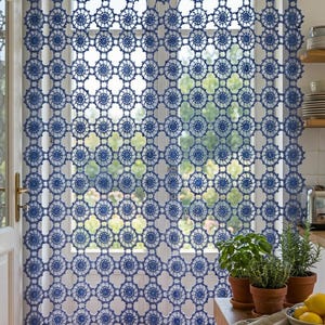 Op de afbeelding: Een blauw gehaakt gordijn met een herhalend cirkelvormig motiefpatroon. Het gordijn hangt aan een horizontale roede voor een raam en toont een ingewikkeld opengewerkt ontwerp waardoor licht door de openingen in de garenstructuur valt.