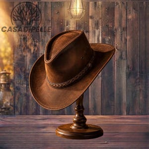 May include: A brown leather cowboy hat featuring a braided leather band around the crown and small ventilation eyelets on the sides. The hat is displayed on a dark wooden stand, showcasing its textured surface and wide, slightly curved brim.