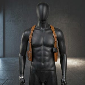 May include: A tan leather shoulder holster system worn on a black mannequin. The harness features adjustable straps with brass-coloured metal buckles, a holster on the left side containing a black handgun, and a magazine pouch on the right side.