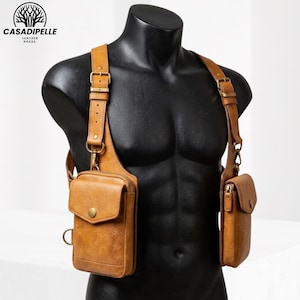 May include: A tan leather shoulder holster bag system featuring two distinct pouches with brass-toned hardware, adjustable buckled straps, and zippered closures. The design includes a larger rectangular pouch on one side and a smaller pouch on the other, both secured with snap buttons, worn over a black torso mannequin.