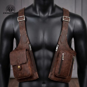 May include: A brown leather shoulder holster bag featuring two symmetrical panels with adjustable silver-tone metal buckles. The right panel includes a flap pocket with a snap button, while the left panel features two vertical zippered compartments.