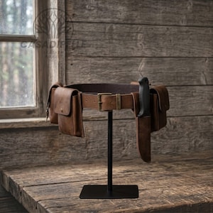 May include: A brown leather utility belt featuring multiple attached pouches with brass-toned metal hardware and a buckle closure. A black-handled knife is secured in a matching leather sheath attached to the belt, which is displayed on a stand.