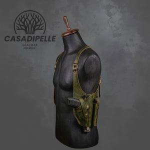 Può includere: Una fondina da spalla in pelle verde oliva scuro con cinghie regolabili, fibbie in metallo e una tasca sagomata per un'arma da fuoco. La fondina è esposta su un busto di manichino nero con il logo CASADIPELLE LEATHER HOUSE visibile.